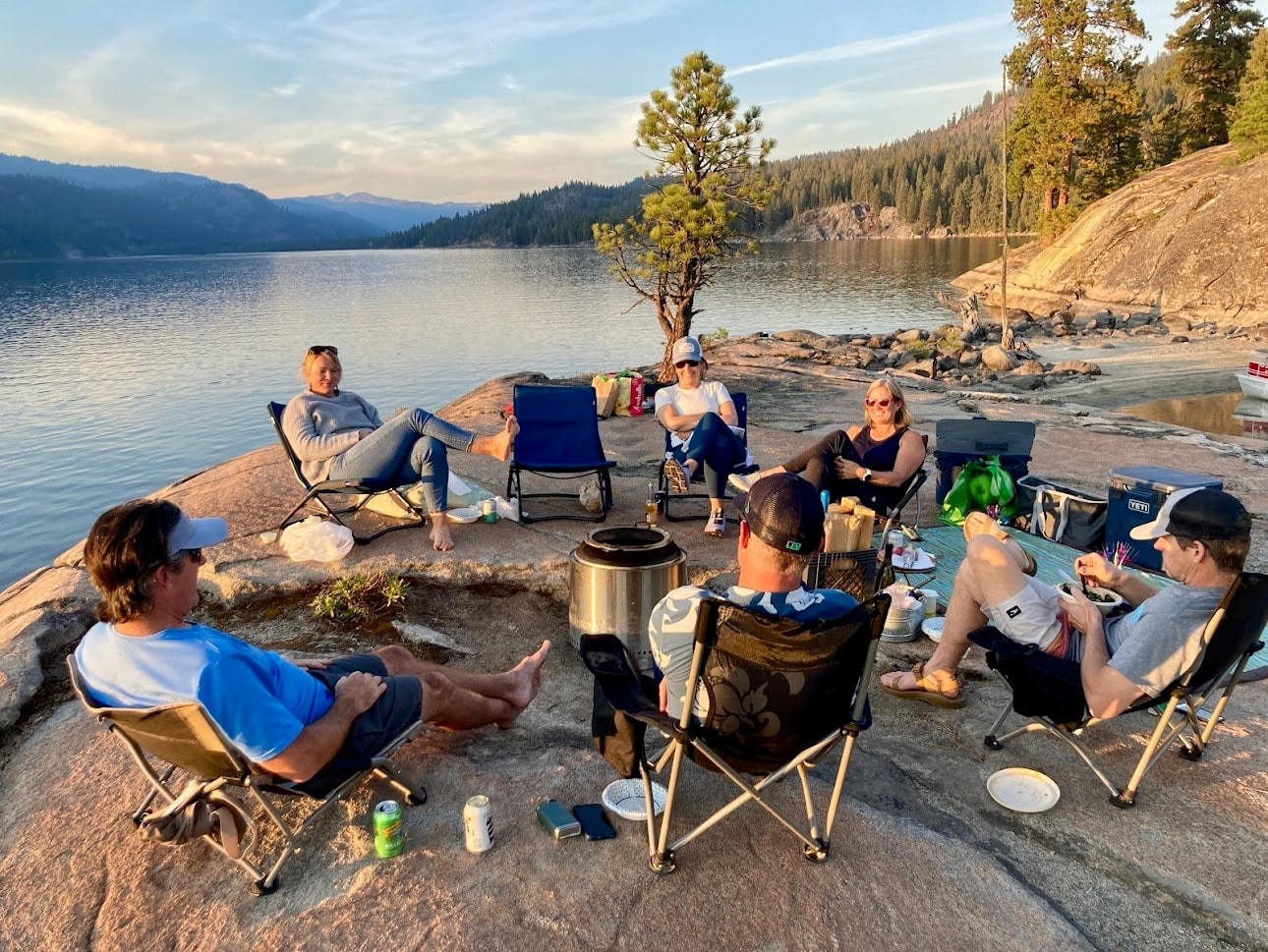 A group of friends gathered in conversation around a lakeside campfire at sunset in McCall, Idaho.